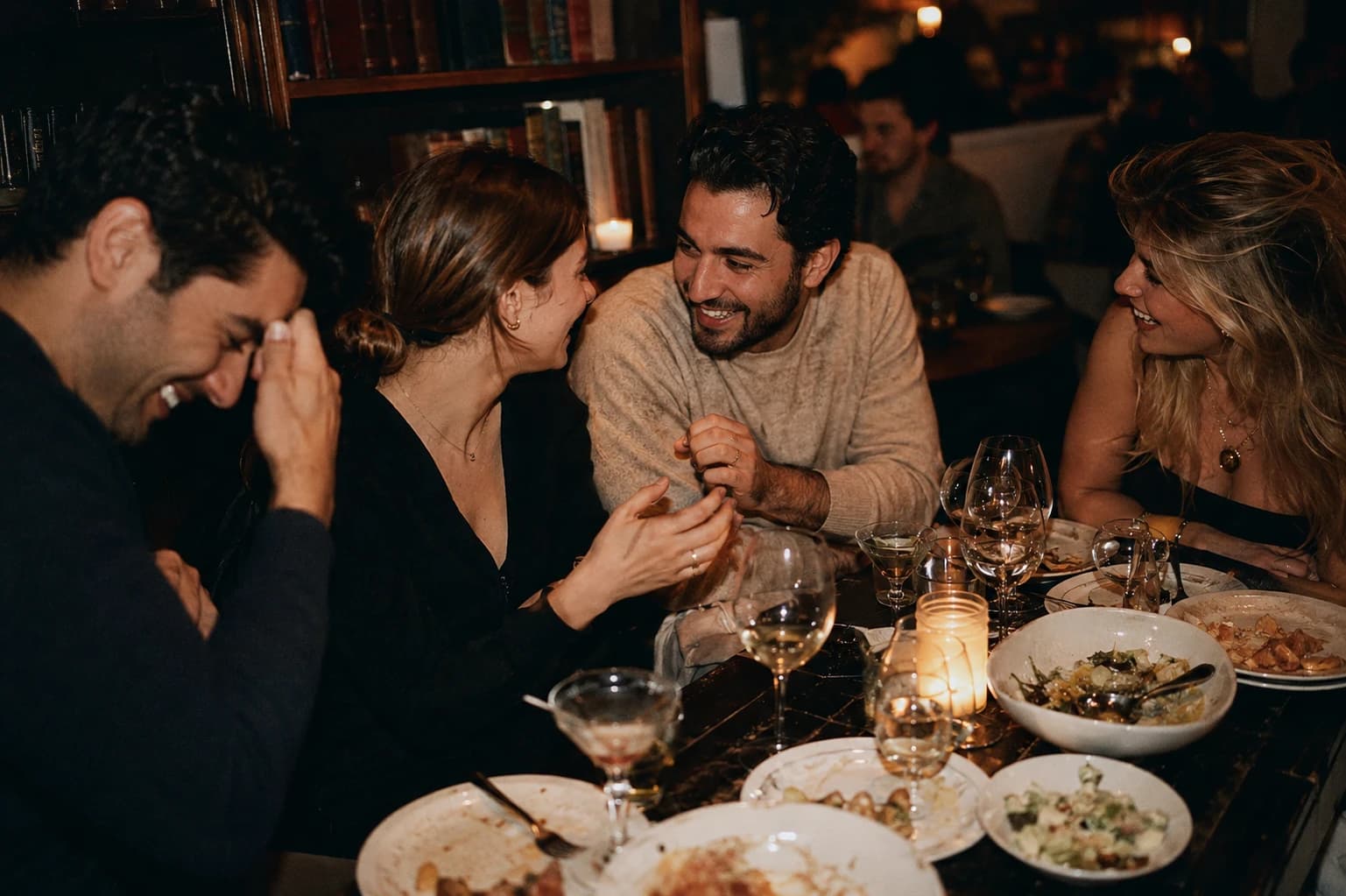 Four friends laughing over a candlelit dinner, half-finished plates and wine glasses on the table