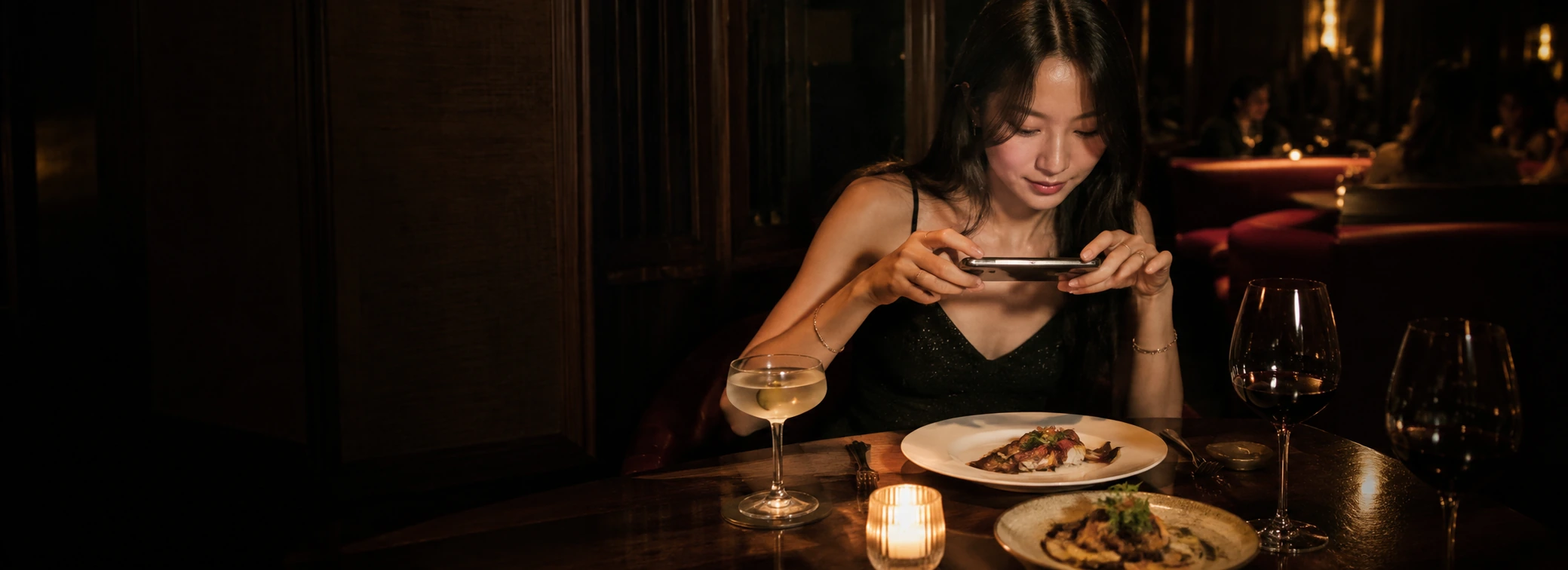 A diner photographing her plates on a phone by candlelight in a dark restaurant